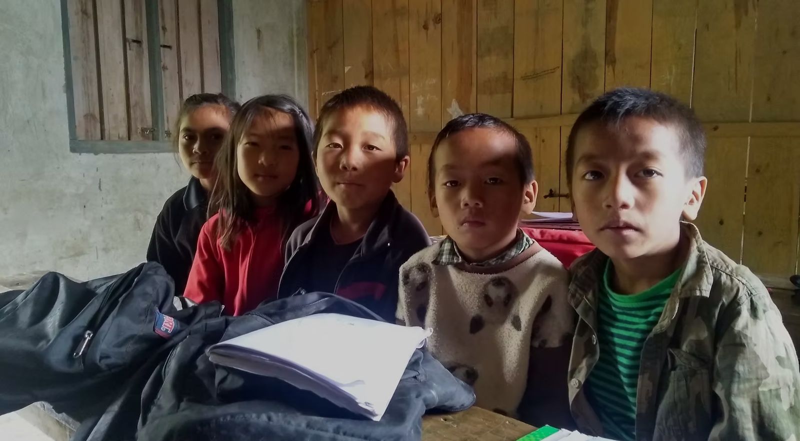 Children sit close together at a wooden desk in a hill school, looking into the camera