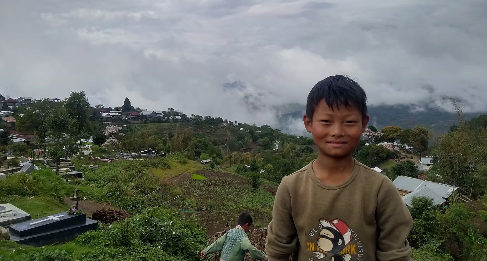A smiling boy stands on a hillside above a village in Ukhrul, clouds over the ridges behind him
